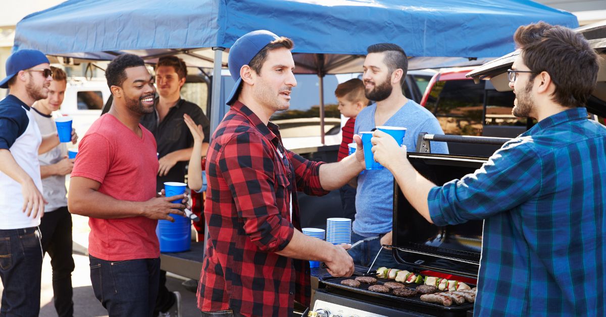 a group of men at a parking lot tailgate wearing sports fan gear, there is also a tent and grill in the background and two men clinking plastic cups
