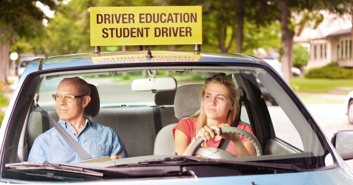 young girl sitting in the driver seat of a small car with her hands on the wheel, a man with a clipboard is in the passenger seat. There is a sign on the roof that says "Driver education student driver"