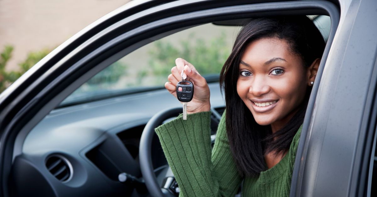 young Black woman wearing a cable-knit sweater and sitting in the driver's seat with the door open, she is holding the car keys and smiling at the camera