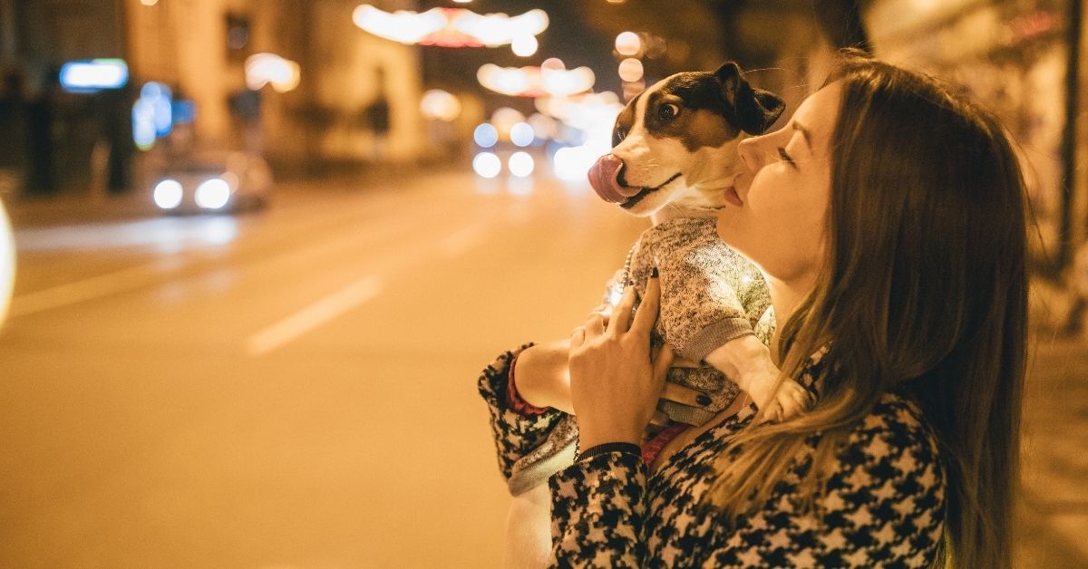 woman holding a dog, standing on a sidewalk under a streelight