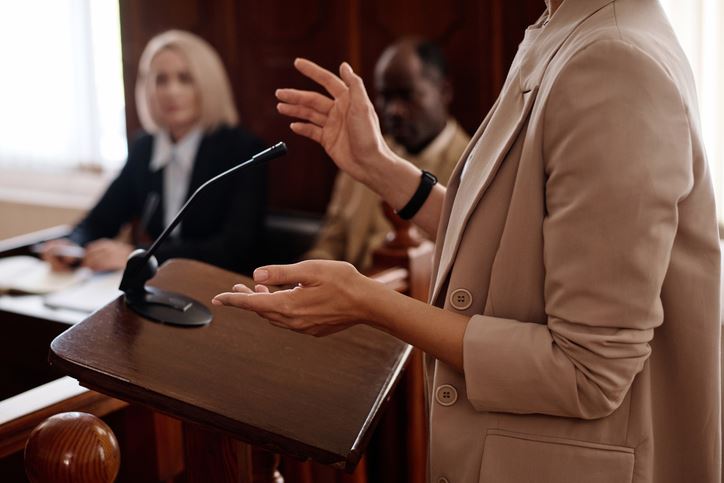 female witness in beige blazer standing by tribune with microphone and testifying in front of jury